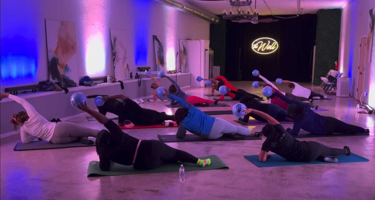 A full Trap Pilates class on mats at The Well, side-plank with the ball raised under purple and blue lights and the glowing Well neon sign